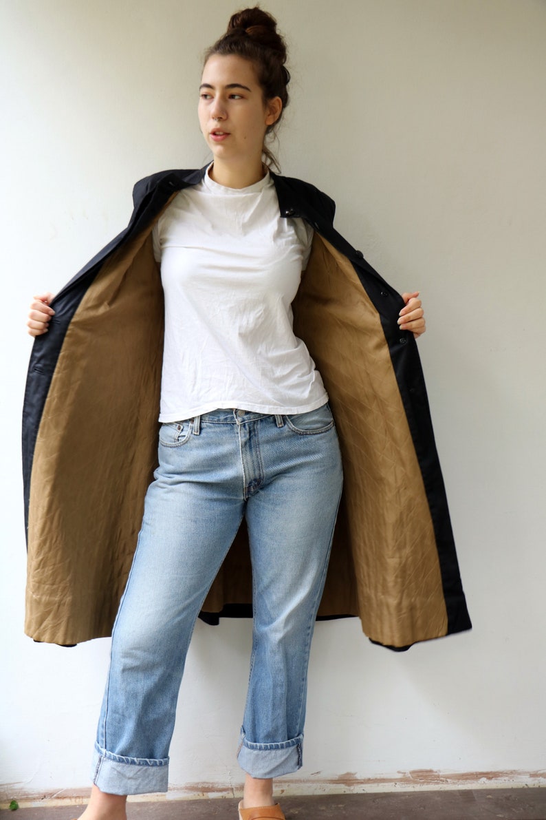 May include: A long black coat with a brown lining. The coat is open and the person wearing it is showing off the lining. The person is wearing blue jeans and a white t-shirt.