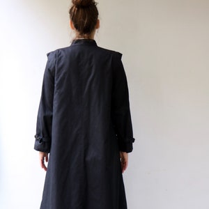 May include: A long black coat with a stand-up collar and a single button fastening. The coat has a slightly flared skirt and is made of a smooth, lightweight fabric.