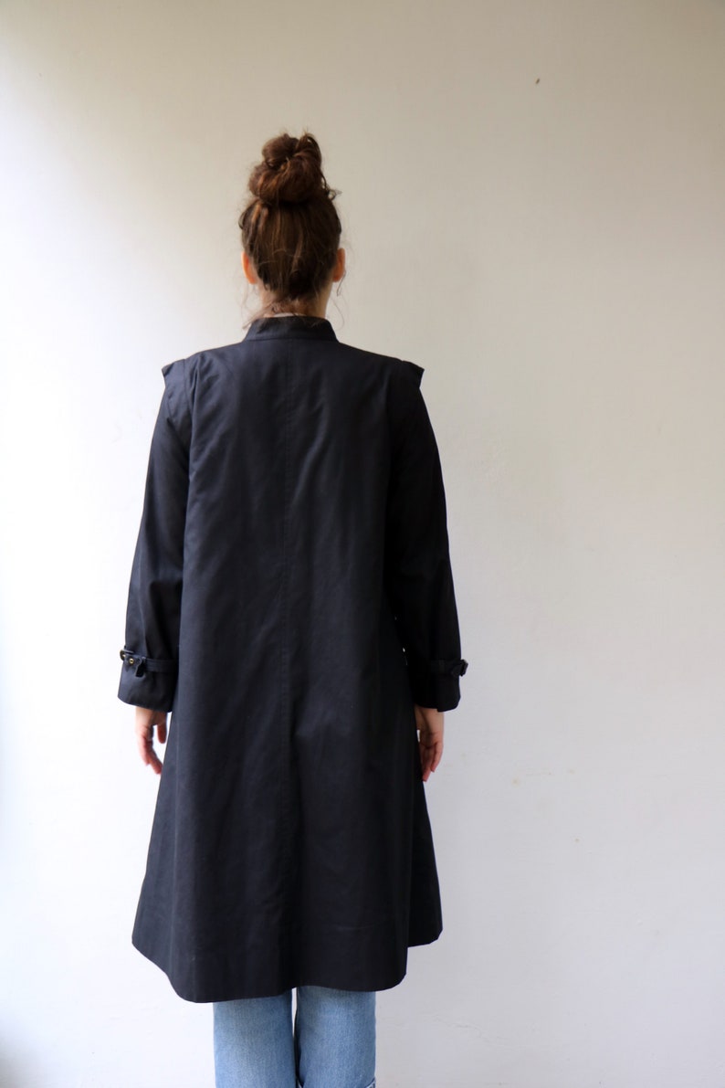 May include: A long black coat with a stand-up collar and a single button fastening. The coat has a slightly flared skirt and is made of a smooth, lightweight fabric.
