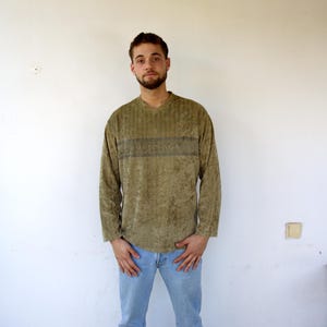 Puede incluir: Un suéter verde texturizado de manga larga con una franja negra y la palabra "VERSACE" impresa en la parte delantera. El suéter lo lleva una persona con el pelo corto castaño y barba. La persona lleva pantalones vaqueros azules.