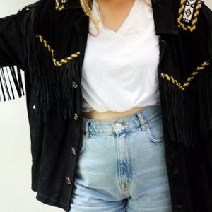May include: A black suede jacket with fringe detailing and yellow embroidery. The jacket is worn over a white t-shirt and blue denim shorts.