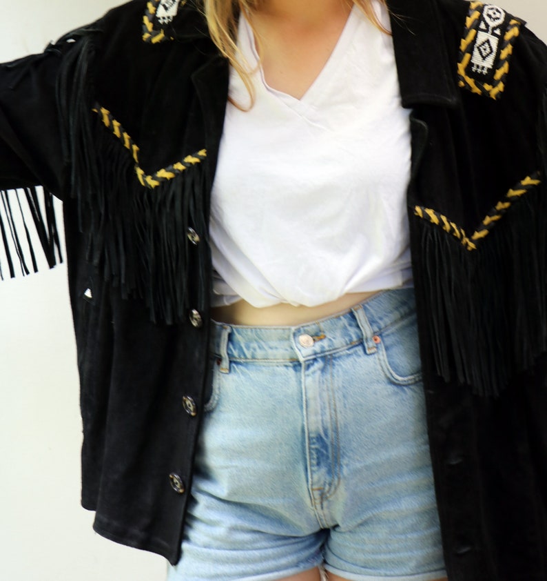 May include: A black suede jacket with fringe detailing and yellow embroidery. The jacket is worn over a white t-shirt and blue denim shorts.