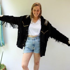 May include: A woman wearing a black suede jacket with fringe and gold embroidery. The jacket has a white t-shirt underneath and blue denim shorts. She is wearing brown leather shoes.