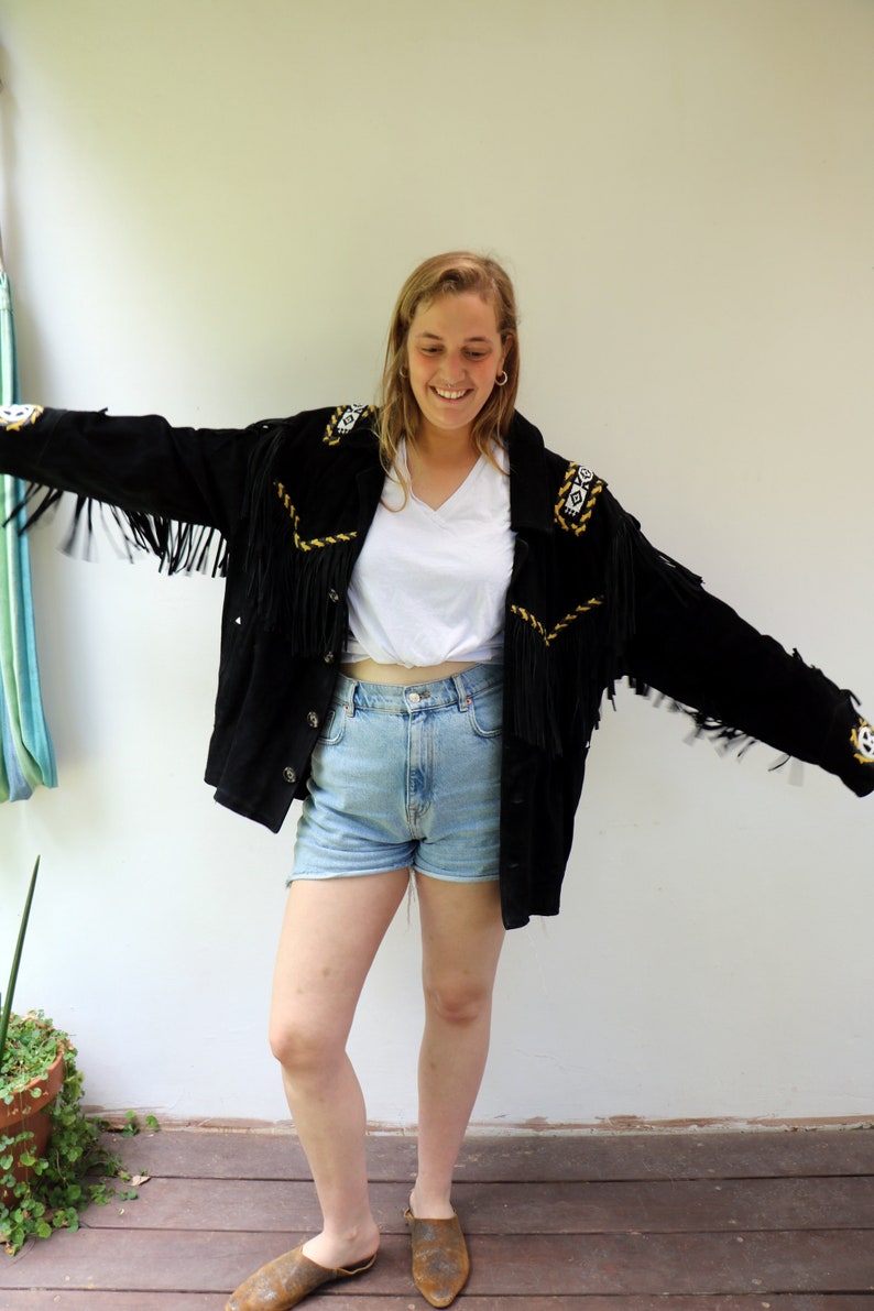 May include: A woman wearing a black suede jacket with fringe and gold embroidery. The jacket has a white t-shirt underneath and blue denim shorts. She is wearing brown leather shoes.
