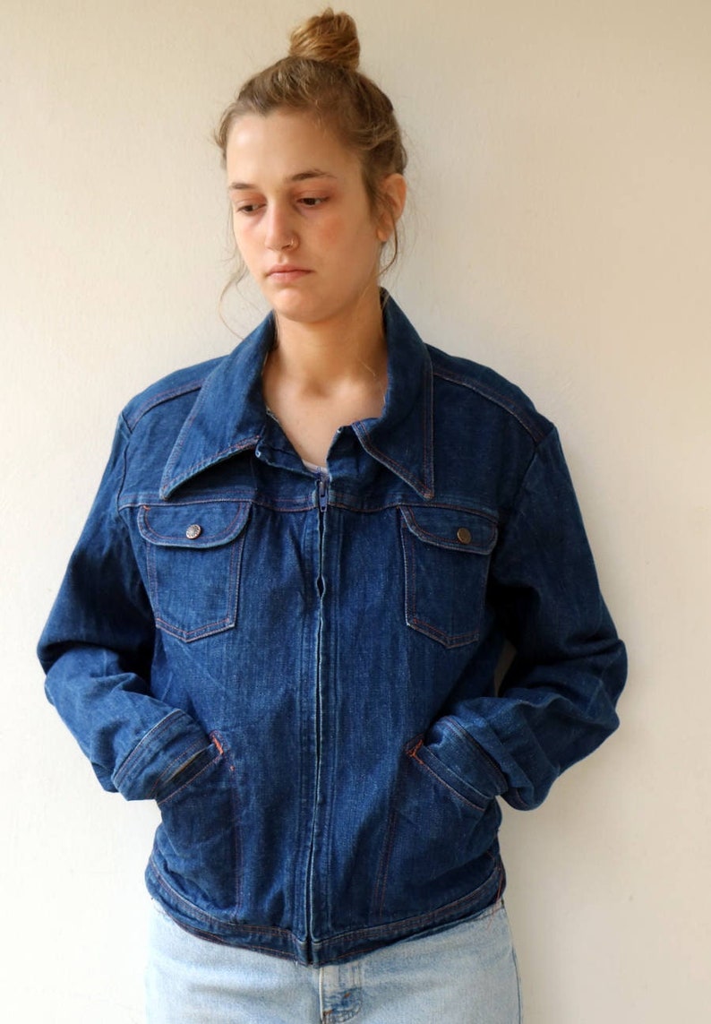May include: A blue denim jacket with a zipper closure and two flap pockets. The jacket has a wide collar and a slightly cropped length.