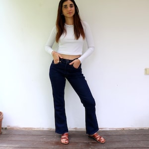 Può includere: Una donna indossa una camicia bianca a maniche lunghe e jeans blu scuro con un leggero svasamento. È in piedi su un pavimento di legno con un muro bianco dietro di lei.