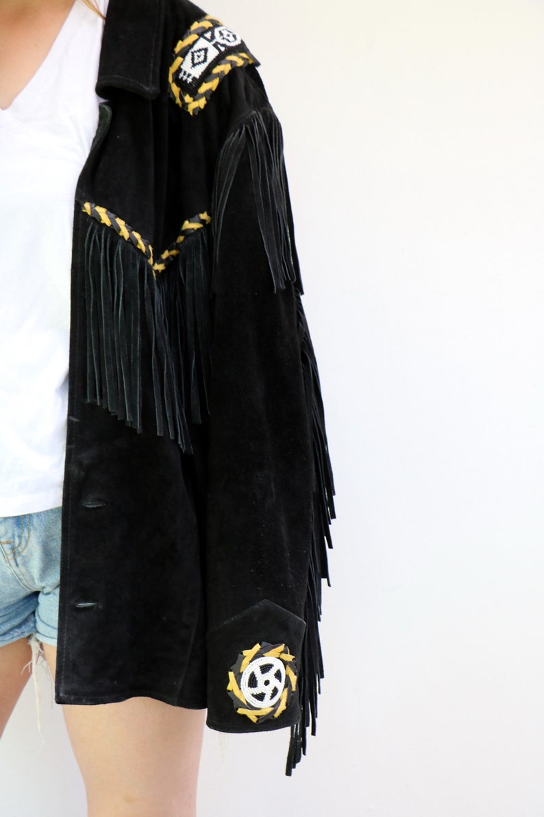 May include: A black suede jacket with fringe detailing and yellow and white embroidered accents. The jacket has a button closure and a collar.