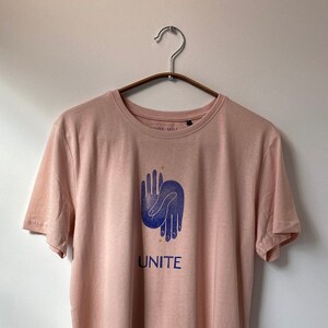 Op de afbeelding: Een lichtroze T-shirt met een blauwe afbeelding van twee handen die in elkaar grijpen. De afbeelding is omringd door kleine gouden sterren. De tekst "UNITE" is onder de afbeelding gedrukt.