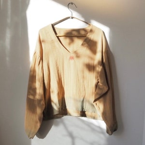 Puede incluir: Una blusa de color marrón claro, de manga larga y cuello en V con un pequeño diseño de sol bordado en la parte delantera. La blusa tiene una manga ligeramente abullonada y un corte holgado.