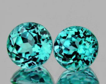May include: Two round, faceted gemstones with a teal color. The gems are displayed on a reflective surface, creating a mirrored effect. The facets catch the light, showcasing the gems' brilliance. The background is a neutral gray.