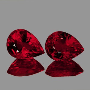 Könnte beinhalten: Zwei birnenförmige rote Edelsteine, wahrscheinlich Rubine, mit einer tiefen, satten Farbe. Die Steine sind facettiert und haben einen hohen Grad an Brillanz.