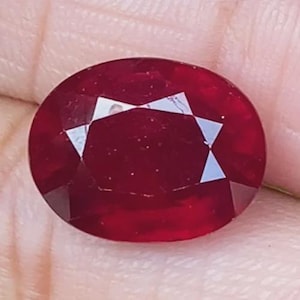 Può includere: Una gemma rosso scuro di forma ovale con una superficie sfaccettata. La pietra presenta una tonalità cremisi brillante e satura e una finitura chiara e lucida con molteplici sfaccettature che riflettono la luce sulla superficie.