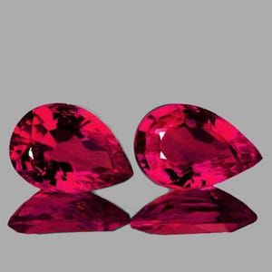 Puede incluir: Dos piedras preciosas en forma de pera, cada una de un color rojo vibrante, con un corte facetado. Las piedras preciosas se muestran sobre un fondo gris.