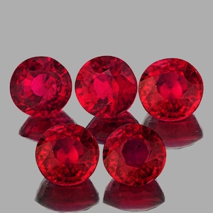Puede incluir: Cinco rubíes redondos y facetados, de color rojo intenso, sobre un fondo gris. Las piedras preciosas tienen estructuras internas y reflejos visibles. Las gemas están dispuestas en un patrón ligeramente descentrado.