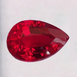 May include: A faceted, pear-shaped ruby gemstone. The gem is a deep, saturated red color, with multiple facets catching the light. The shape is a classic teardrop, perfect for jewelry.