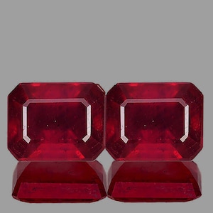 May include: Two vibrant red emerald-cut gemstones, each with a polished, faceted surface. The stones are set against a gray background, showcasing their brilliance and color.