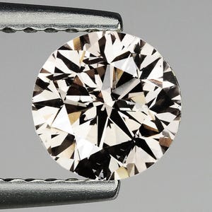 May include: A round-cut diamond held by tweezers. The gemstone has a complex, multifaceted design with a mix of light and dark facets, creating a sparkling effect. The diamond's color is a light champagne hue, and it is set against a neutral gray background.