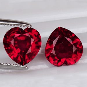 Puede incluir: Dos gemas rojas en forma de corazón, facetadas. Una es sostenida por unas pinzas plateadas. Las gemas tienen un corte brillante, reflejando la luz. El fondo es blanco.