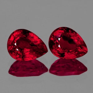 Puede incluir: Dos piedras preciosas rojas en forma de pera con un corte facetado. Las piedras preciosas están colocadas sobre un fondo gris.
