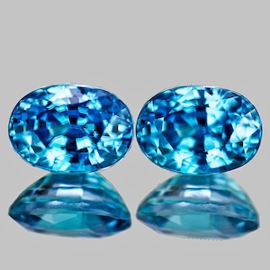 May include: Two oval-shaped blue gemstones with a faceted cut. The gemstones are set against a gray background.