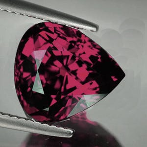 Puede incluir: Una gema facetada en forma de pera, de un tono magenta intenso, que transiciona a negro en algunas áreas. La gema es sostenida por unas pinzas plateadas, mostrando su intrincada estructura interna y superficies reflectantes. El corte revela patrones geométricos.