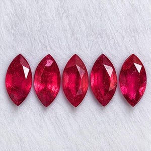 May include: Five loose marquise-cut gemstones in a vibrant, deep red colour. Each stone features a pointed, elongated oval shape with multiple facets that reflect light, creating a brilliant, sparkling appearance across the surface of the gems.