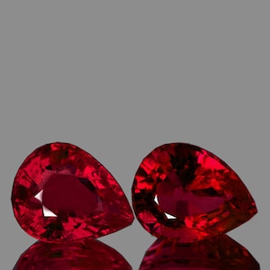 May include: Two pear-shaped red gemstones with a faceted cut. The gemstones are set against a gray background.