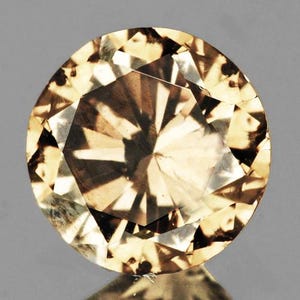 May include: A round-cut, light brown gemstone with multiple facets. The gemstone has a reflective surface and is set against a gray background. The facets create a starburst pattern in the center.