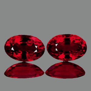 May include: Two oval-shaped, deep red gemstones with a high polish. The gemstones are set against a grey background.