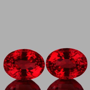 May include: Two oval-cut, faceted gemstones with a deep red color. The gems are displayed against a gray background, with a reflective surface below them. The stones have a brilliant, sparkling appearance.