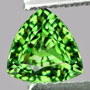 Op de afbeelding: Een gefacetteerde, driehoekige edelsteen met een levendige, heldergroene kleur. De edelsteen wordt vastgehouden door een zilveren metalen gereedschap, dat de ingewikkelde slijping en helderheid benadrukt. De facetten van de edelsteen reflecteren licht, waardoor een sprankelend effect ontstaat.