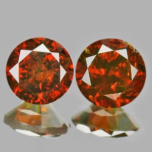 May include: Two round, faceted gemstones with a fiery orange-red hue. Each gem features a central white geometric design. The gems are displayed on a reflective surface, showcasing their brilliance and clarity. The gems are likely for jewelry.