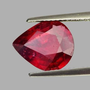 Può includere: Una pietra preziosa di forma a pera di colore rosso intenso, tenuta da una pinza. La pietra preziosa è sfaccettata e ha un'alta lucentezza.