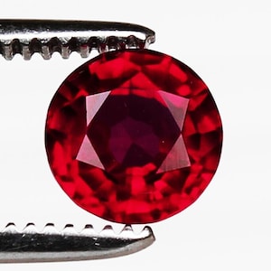 Puede incluir: Una gema facetada, redonda, de color rojo intenso, sujeta por una herramienta plateada. La gema tiene un tallado brillante con múltiples facetas que reflejan la luz, creando un efecto brillante. La gema es de un color rojo vibrante y saturado.