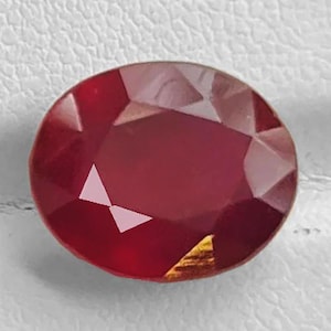 May include: A deep red, oval-cut gemstone with a faceted surface. The stone displays a rich, saturated crimson hue with multiple geometric facets that reflect light, creating a polished and brilliant appearance across its entire surface.