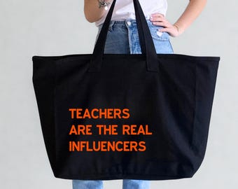 May include: Black tote bag with the orange text "TEACHERS ARE THE REAL INFLUENCERS." The bag has two long handles and is held by a person wearing a white t-shirt and blue jeans.