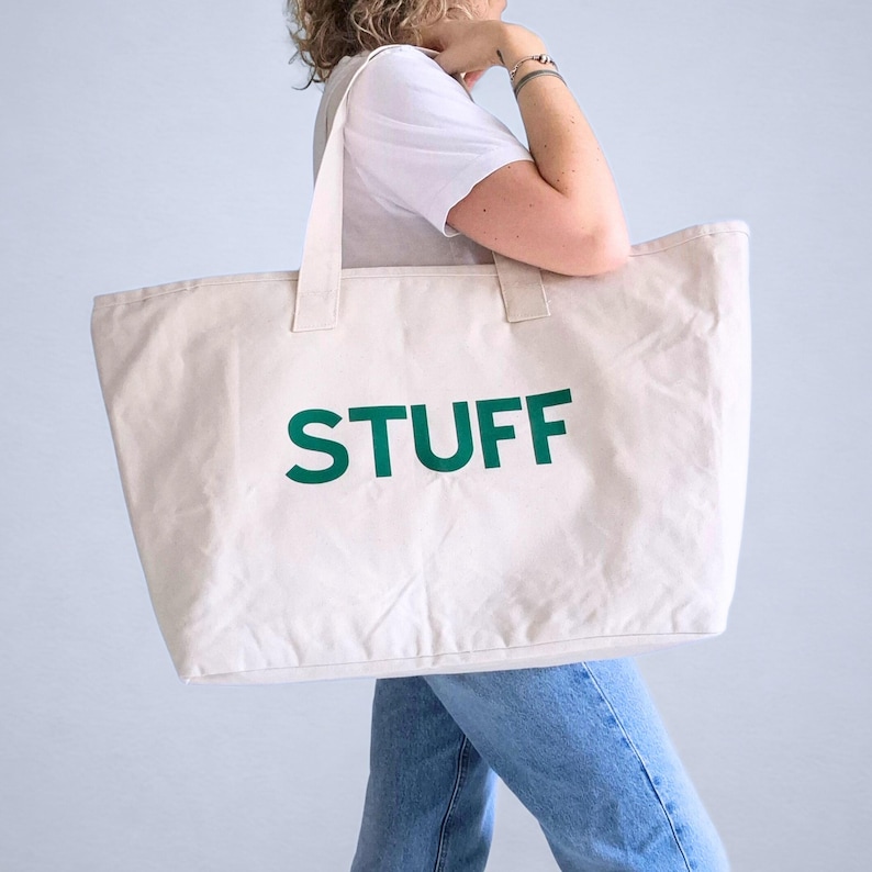May include: A large, cream-colored tote bag with the word "STUFF" printed in bold, dark green letters. The bag has two sturdy handles and is carried over the shoulder. The bag is made of a durable fabric.