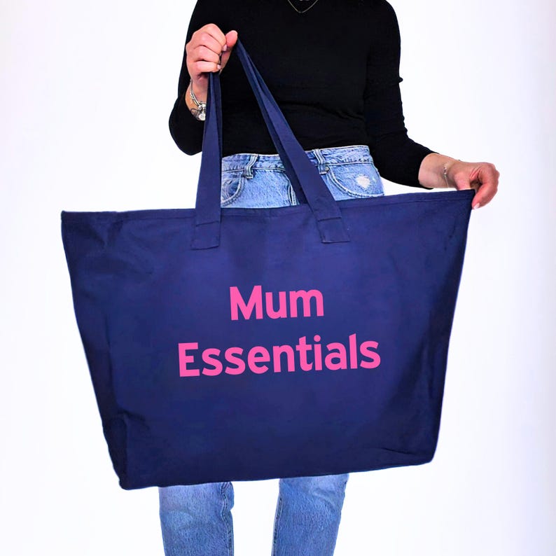 May include: A navy blue tote bag with the words "Mum Essentials" in pink. The bag has two long handles and is held up by a person. The bag is made of a sturdy material and is designed for carrying items.