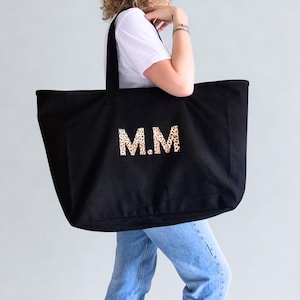 Puede incluir: Bolso tote negro con las iniciales "M.M" en diseño de estampado de leopardo. El bolso tiene dos asas negras. La persona que sostiene el bolso lleva una camiseta blanca y vaqueros azules.