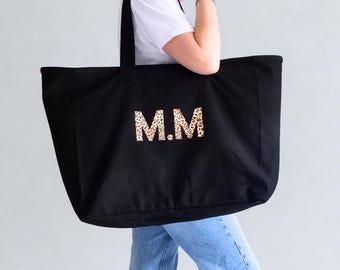 May include: Black tote bag with the initials "M.M" in a leopard print design. The bag has two black handles and is being held by a person wearing a white t-shirt and blue jeans.
