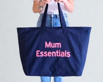 May include: A navy blue tote bag with the words "Mum Essentials" printed in bright pink. The bag has two handles and is held up by a person. The bag is made of a sturdy material and is designed for carrying items.