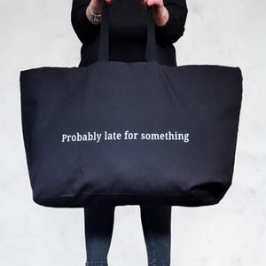 Puede incluir: Una gran bolsa de lona negra con el texto "Probably late for something" impreso en blanco.