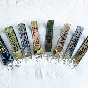 May include: A collection of personalized keychains with various names embroidered on camouflage fabric. Each keychain features a different name in a contrasting color, with a small bow detail and a silver-tone metal clasp. The keychains are arranged on a white surface.