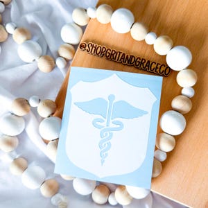 May include: A light blue sticker featuring a white medical caduceus symbol, set against a light blue shield shape. The sticker is on a wooden surface, surrounded by a garland of white and natural wooden beads. The text "@SHOPGRITANDGRACECO" is visible.