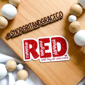 May include: A red sticker with the word "RED" in large, bold letters, with the phrases "remember everyone deployed" and "until they all come home." A wooden sign with the text "@SHOPGRITANDGRACECO" is also present.