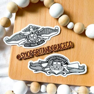 May include: Three stickers on a wooden surface. The stickers feature black and white illustrations of military emblems and text. One sticker reads "FLEET MARINE FORCE", another reads "EXPEDITIONARY", and a third reads "@SHOPGRITANDGRACECO". Decorative wooden beads are in the background.