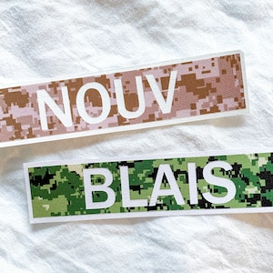 May include: Two rectangular stickers. The top sticker has a pink and brown pixelated camouflage pattern with the word "NOUV" in white block letters. The bottom sticker has a green and black pixelated camouflage pattern with the word "BLAIS" in white block letters.