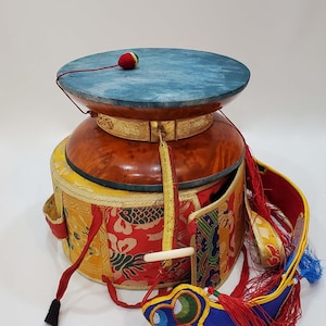 Puede incluir: Un molinillo de oración tibetano tradicional con una parte superior azul, un cuerpo de madera marrón y cubiertas de tela de colores. La rueda está decorada con ribetes dorados y tiene un borla roja.