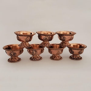 May include: Six small copper bowls with intricate designs. The bowls have a rounded base and a flared lip. They are arranged in a row on a white surface.