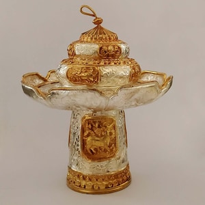 Op de afbeelding: Een decoratieve standaard van zilver en goud met een lotusvormige basis en een meerlagig bovendeel. De standaard is versierd met ingewikkelde bloem- en diermotieven.